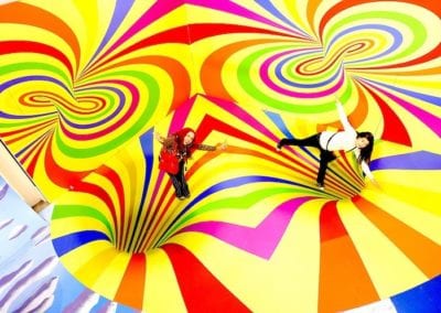 An aerial shot of two happy humans standing on a bright yellow floor painted with swirls to make it appear like it has swirly vortexes.